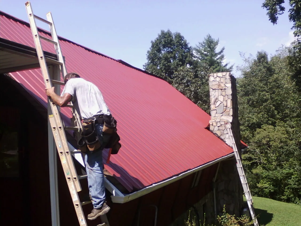 Roofer on a ladder beside a red metal roof and stone chimney — Soffit & Fascia Repair by Roofing Brothers in Calder